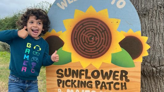 an excited child stands by the sign at the entrance of the maize with thumbs up